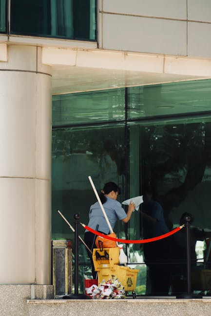 A person cleaning a large glass window from the outside during daytime, holding a squeegee and wearing a dark jacket with a scarf. The window has visible smudges, dust, and water spots, indicating it 