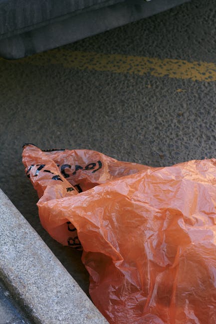 A pile of discarded bulky items and trash bags on asphalt ground next to a stone wall, under a clear blue sky with overhead power lines. Visible objects include a large, worn inflatable tire, a yellow