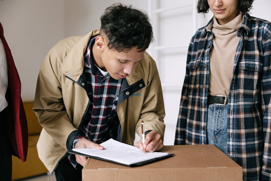 A person in a beige jacket and plaid shirt is signing a document or checklist on a clipboard placed on top of a cardboard box in an indoor setting. To the right, another individual in a blue and brown plaid shirt, beige turtleneck, and jeans is observing. The background includes white shelving units and a window, providing natural lighting. The scene appears to relate to a residential or commercial cleaning process, aligning with surface cleaning or deep cleaning activities associated with end-of-tenancy preparation, as highlighted on the Merton Cleaners website for Wimbledon SW19. The environment looks clean and organized, emphasizing the professional approach of Merton Cleaners in hygiene and maintenance tasks.