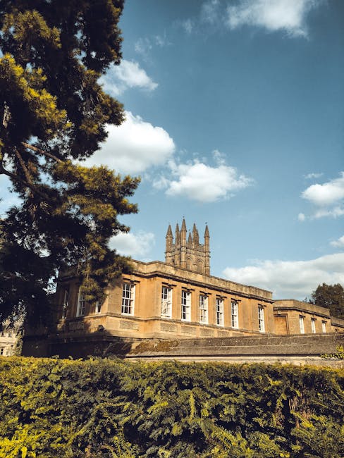 An exterior view of a historic stone building featuring large rectangular windows with white frames and decorative moldings, set amidst lush greenery and tall trees. The building has a Renaissance architectural style with detailed stonework and a prominent tower with spires at the top. The sky above is bright blue with scattered white clouds, and the foreground includes well-maintained bushes and grass, highlighting a clean and orderly landscape. This scene exemplifies the importance of surface cleaning and maintenance for historic structures, aligning with professional deep cleaning and sanitisation practices provided by Merton Cleaners, as referenced in the Merton end-of-tenancy clean: SW19 (Wimbledon) checklist.