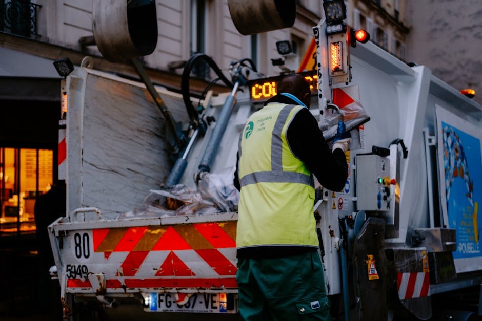 Two individuals wearing white gloves and casual clothing are engaged in waste collection outdoors, gathering and tying up a large beige trash bag filled with rubbish. The scene takes place in a natura