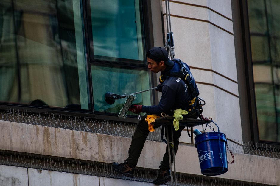A professional cleaning worker wearing a safety helmet is performing surface cleaning on the exterior glass facade of a modern commercial building. The worker is seated on a platform secured with harn