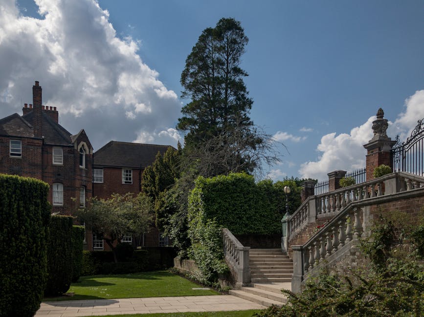 A view of a residential property with a brick mansion-style house featuring multiple chimneys and white-framed windows, set amidst well-maintained gardens with tall, trimmed hedges and lush green gras